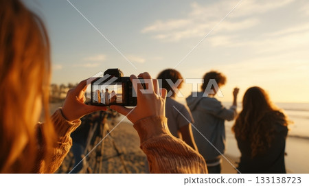 Girl photographing friends sunset beach scene with warm golden light, capturing joyful summer memories and friendship moments during a seaside evening adventure. 133138723