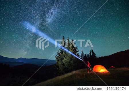 Man hiker stands under dazzling starry sky, shining flashlight into heavens. Milky Way stretches above, glowing tent nearby, creating magical and serene scene in peaceful mountain wilderness. 133139474