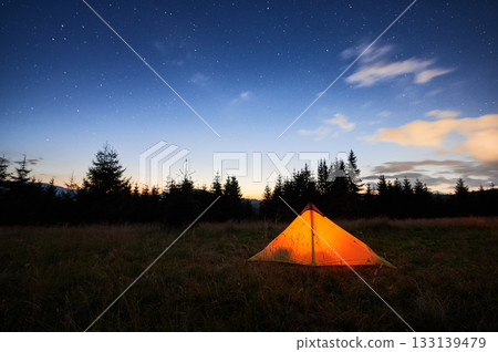 Glowing orange tent on grassy field under starry sky, with hint of dawn's light on horizon. Surrounded by pine trees, tranquil scene captures serene beauty of early morning in nature. Glowing orange tent on grassy field under starry sky, with hint of dawn's light on horizon. Surrounded by pine trees, tranquil scene captures serene beauty of early morning in nature. 133139479