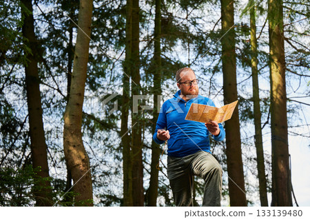 Man traveler in glasses stands in dense forest, holding map in one hand and compass in other. Tourist planning outdoor adventure or hiking route, amidst tall trees and natural surroundings. Man traveler in glasses stands in dense forest, holding map in one hand and compass in other. Tourist planning outdoor adventure or hiking route, amidst tall trees and natural surroundings. 133139480