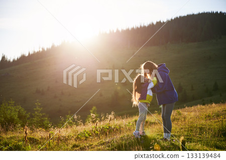 Young woman tourist spending time together with little daughter in sunlit meadow, against stunning mountain backdrop at sunset. Joyful moment captures essence of carefree fun and natural beauty. Young woman tourist spending time together with little daughter in sunlit meadow, against stunning mountain backdrop at sunset. Joyful moment captures essence of carefree fun and natural beauty. 133139484