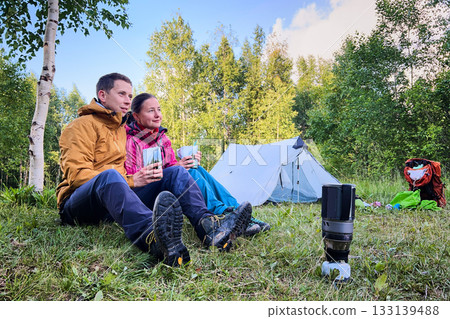 Couple sits on grass near tent in lush forest, enjoying meals from packaged pouches. Dressed in outdoor gear, with portable camping stove and backpack nearby. Green trees and foliage on background. Couple sits on grass near tent in lush forest, enjoying meals from packaged pouches. Dressed in outdoor gear, with portable camping stove and backpack nearby. Green trees and foliage on background. 133139488
