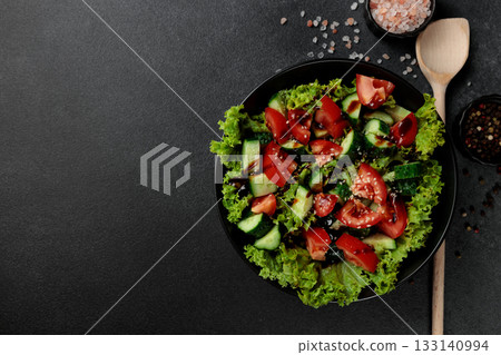 Vegetable salad of tomatoes, cucumbers, onions, lettuce dressed with aromatic dressing in a black plate on a gray background Vegetable salad of tomatoes, cucumbers, onions, lettuce dressed with aromatic dressing in a black plate on a gray background 133140994