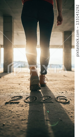 A person in jeans and brown boots walks away towards bright sunlight in a desolate concrete structure, with '2026' written on the dusty ground. A person in jeans and brown boots walks away towards bright sunlight in a desolate concrete structure, with '2026' written on the dusty ground. 133143292
