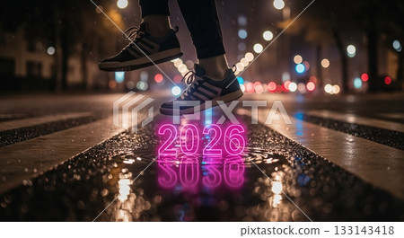 A person's feet in sneakers are captured mid-air above a glowing neon 2026 reflected in a puddle on a wet city street at night with bokeh lights. 133143418