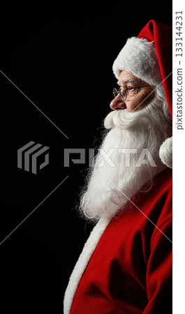 An elderly man portraying Santa Claus, wearing a red suit and hat with a long white beard and spectacles, looks left against a dramatic dark background. An elderly man portraying Santa Claus, wearing a red suit and hat with a long white beard and spectacles, looks left against a dramatic dark background. 133144231