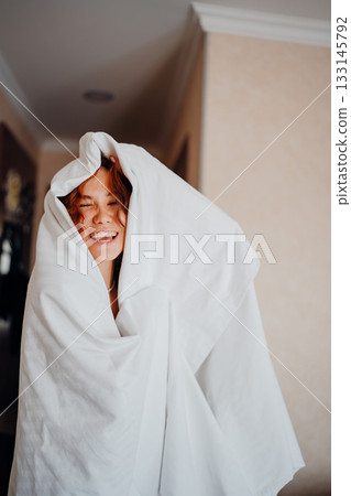 Young woman playfully wrapped in a white blanket inside a cozy room 133145792