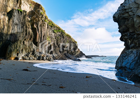 Waves crash onto the dark sand beach at Sheep Cove in Ireland, with steep, rocky cliffs rising up on either side under a partly cloudy, blue sky. Waves crash onto the dark sand beach at Sheep Cove in Ireland, with steep, rocky cliffs rising up on either side under a partly cloudy, blue sky. 133146241