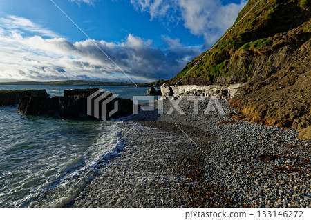 Waves crash on a rocky beach in Ireland, with a cliff in the background. 133146272