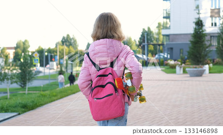 Rear view of girl walking to school in the morning carrying a vibrant skateboard and wearing a pink schoolbag Rear view of girl walking to school in the morning carrying a vibrant skateboard and wearing a pink schoolbag 133146898