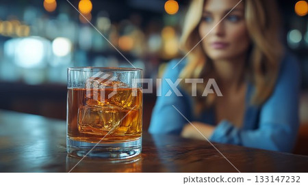 Glass of whiskey with ice cubes on wooden bar counter with defocused woman in background, capturing evening drink atmosphere in modern setting Glass of whiskey with ice cubes on wooden bar counter with defocused woman in background, capturing evening drink atmosphere in modern setting 133147232