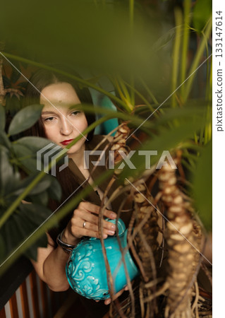 Focused female gardener holding jug among lush indoor foliage Focused female gardener holding jug among lush indoor foliage 133147614