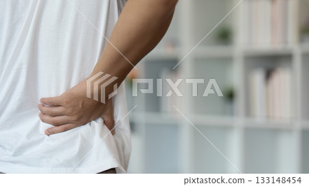 Back Pain Awareness. Young man holding lower back in discomfort while seated. Back Pain Awareness. Young man holding lower back in discomfort while seated. 133148454