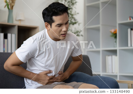 Abdominal Pain. Young man experiencing discomfort while sitting at home. 133148490