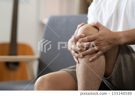 Knee Pain Relief. Man holding knee in discomfort while sitting on couch. Knee Pain Relief. Man holding knee in discomfort while sitting on couch. 133148580