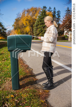 Woman standing beside a roadside mailbox on a sunny autumn day, holding an envelope and preparing to place it inside while colorful trees line the street. 133148618