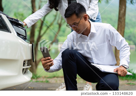 Insurance Assessment. A man inspecting a car while using a smartphone for insurance evaluation. 133148620