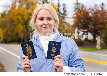 Middle-aged woman standing outdoors in autumn, holding a Canadian passport in one hand and a United States passport in the other near a road. Middle-aged woman standing outdoors in autumn, holding a Canadian passport in one hand and a United States passport in the other near a road. 133148623