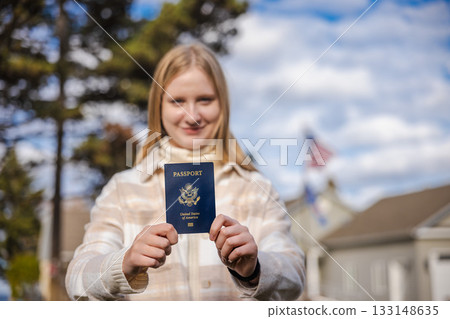 Teenage girl standing outdoors and holding a blue US passport in both hands, with houses and an American flag visible in the blurred background. Teenage girl standing outdoors and holding a blue US passport in both hands, with houses and an American flag visible in the blurred background. 133148635
