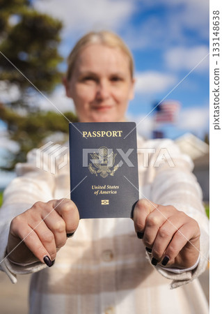 Woman holding a United States passport at arm's length toward the camera on a sunny day, with a blurred American flag and suburban houses in the background. 133148638