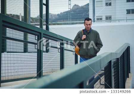 Man With Green Jacket and Orange Backpack Walking Across an Urban Ramp 133149266