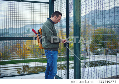 Man With Backpack Locks Gate On Fenced Balcony Overlooking Cityscape Urban View Man With Backpack Locks Gate On Fenced Balcony Overlooking Cityscape Urban View 133149271