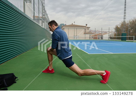 Athletic Man Performs Lunge Stretch On Outdoor Tennis Court Preparing For Workout Session Athletic Man Performs Lunge Stretch On Outdoor Tennis Court Preparing For Workout Session 133149278