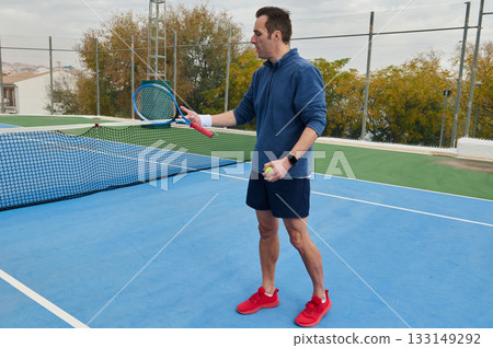 Man In Blue Hoodie Plays Tennis On Outdoor Court With Ball And Racket 133149292
