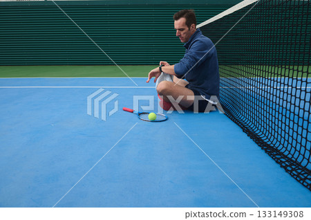 Man Sits on Tennis Court Near Net With Racket And Ball, Resting After Match Man Sits on Tennis Court Near Net With Racket And Ball, Resting After Match 133149308