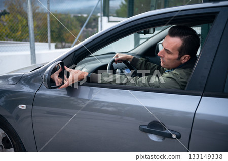 Focused Man Driving a Car Reaching for Side Mirror Inside a Modern Gray Vehicle 133149338
