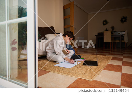 Teen Boy Studying At Home On A Rug With Laptop And Notebooks, Focused And Cozy 133149464