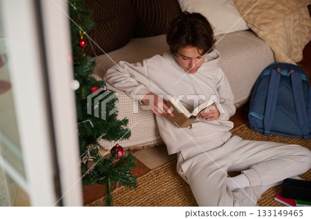 Teen Boy Reading a Book by Christmas Tree at Home With Backpack Nearby 133149465