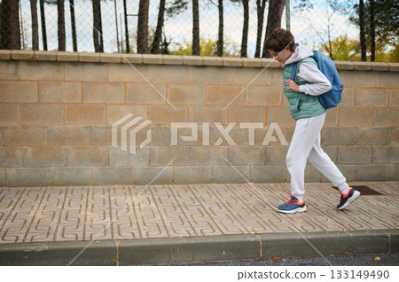 Teen Boy Walking With Backpack Along City Sidewalk Beside A Brick Wall 133149490