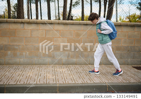 Teen Boy Walking With Backpack On Sidewalk In Casual Winter Outfit Near A Brick Wall 133149491