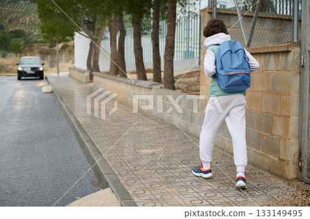 Teenager With Blue Backpack Walking Along Sidewalk On Suburban Street Teenager With Blue Backpack Walking Along Sidewalk On Suburban Street 133149495