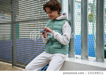Teenage Boy Using Smartphone While Sitting by Fence in Casual Outdoor Setting Teenage Boy Using Smartphone While Sitting by Fence in Casual Outdoor Setting 133149511