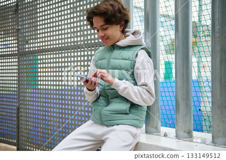 Teen Boy Using Smartphone At Outdoor Sports Field Behind Metal Fence, Casual Vest Teen Boy Using Smartphone At Outdoor Sports Field Behind Metal Fence, Casual Vest 133149512
