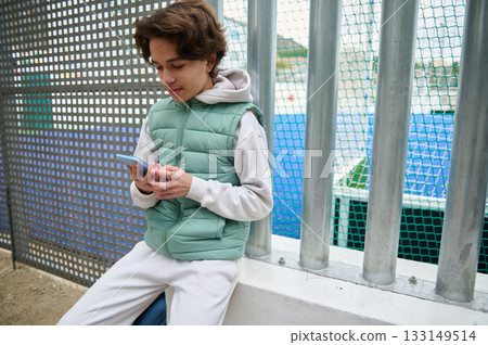 Teenage Boy Using Smartphone By Fence Near Outdoor Court In Green Puffer Vest 133149514