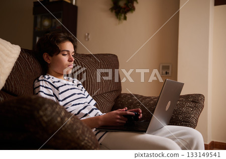 Teenage Boy Relaxing At Home With Laptop And Game Controller On Couch Teenage Boy Relaxing At Home With Laptop And Game Controller On Couch 133149541