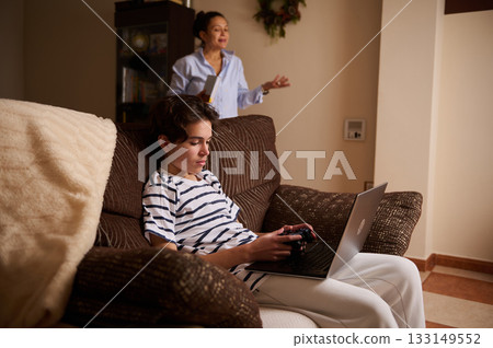 Teen Gamer On Laptop On Couch With Mom Talking In Background At Home Teen Gamer On Laptop On Couch With Mom Talking In Background At Home 133149552