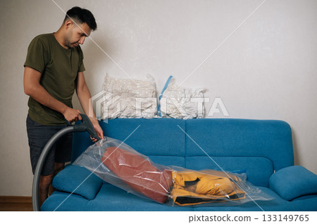 Side view of man using vacuum cleaner to remove air from vacuum storage bag with clothes and placing it on sofa in living room, organizing seasonal wardrobe for winter. Concept of household chores. Side view of man using vacuum cleaner to remove air from vacuum storage bag with clothes and placing it on sofa in living room, organizing seasonal wardrobe for winter. Concept of household chores. 133149765