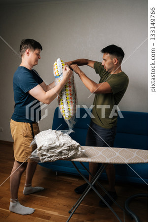 Vertical shot of two men using vacuum storage bags to compress bedding and pillows, optimizing space and making moving or storing household items easier. Concept of household chores and housework. Vertical shot of two men using vacuum storage bags to compress bedding and pillows, optimizing space and making moving or storing household items easier. Concept of household chores and housework. 133149766