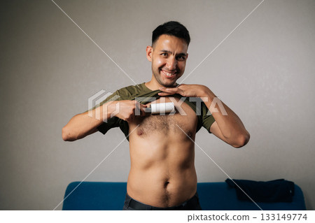 Portrait of happy excited young man lifting shirt and using sticky lint roller to remove hair and fluff from chest at home, showcasing humorous approach to personal grooming, smiling looking camera. 133149774