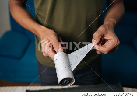 Man carefully peeling off used layer of sticky lint roller, preparing to clean clothes from dust, hair and fibers, ensuring garments remain tidy and fresh. Concept of household chores and housework. 133149775