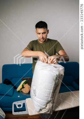 Vertical shot of focused man closing large transparent vacuum storage bag filled with white bedding, using double-zip closure to create airtight seal for optimal space efficiency and protection. 133149776