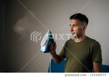 Portrait of handsome young man holding modern iron while looking at cloud of steam rising as smoothly presses wrinkles from clothes in tidy domestic setting. Concept of household chores 133149777
