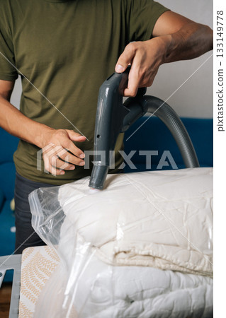 Close-up shot of unrecognizable man using vacuum cleaner to extract air from storage bag filled with bedding, positioned on ironing board, creating efficient space-saving storage solution.  133149778