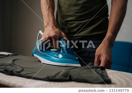 Unrecognizable man ironing dark green t-shirt on ironing board, using steam iron to remove wrinkles and achieve crisp at home, close-up. Concept of household chores and housework. Unrecognizable man ironing dark green t-shirt on ironing board, using steam iron to remove wrinkles and achieve crisp at home, close-up. Concept of household chores and housework. 133149779