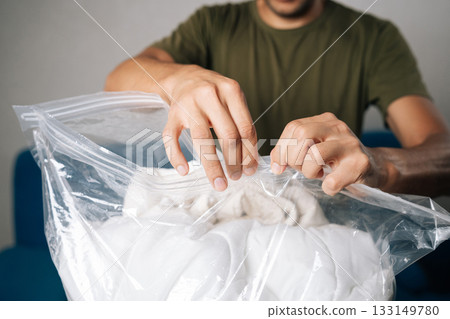 Close-up male hands closing large transparent vacuum storage bag filled with white bedding, using double-zip closure to create airtight seal for optimal space efficiency and protection. 133149780