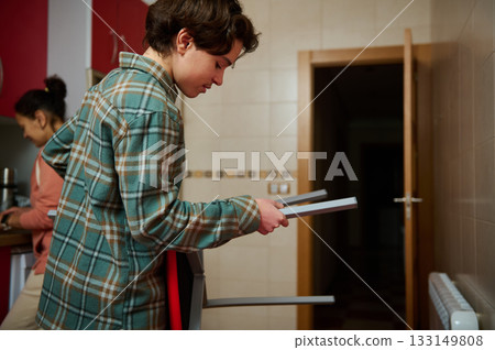 Teenage Boy In Plaid Shirt Examines Furniture in Kitchen While Family Works Nearby 133149808